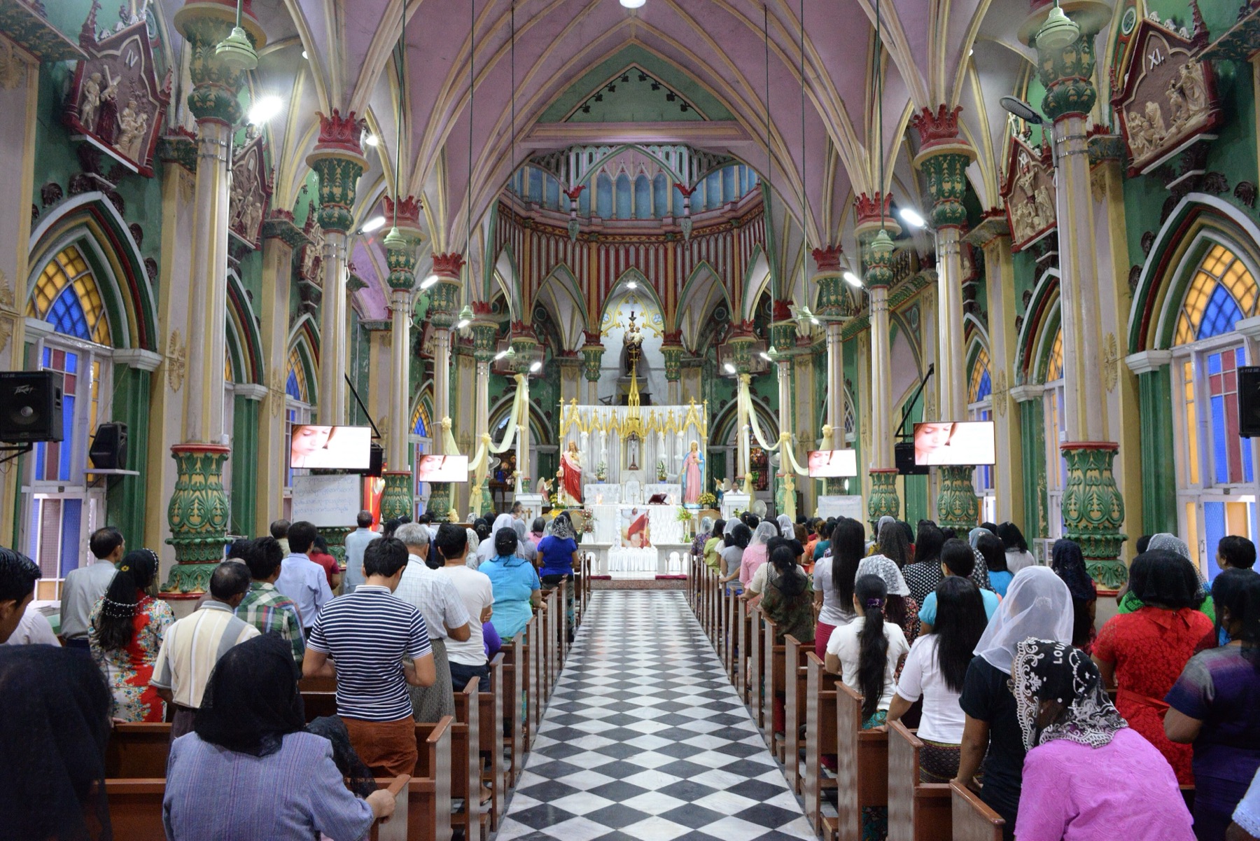 A full congregation stands in St. Joseph's Church in Mandalay during Mass, facing a brightly decorated altar.
